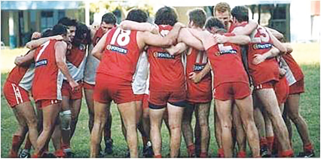 A retro image of a group of ANZA Wombats players in red and white uniforms huddle together on a grassy field, arms linked in a pre-game or halftime circle.