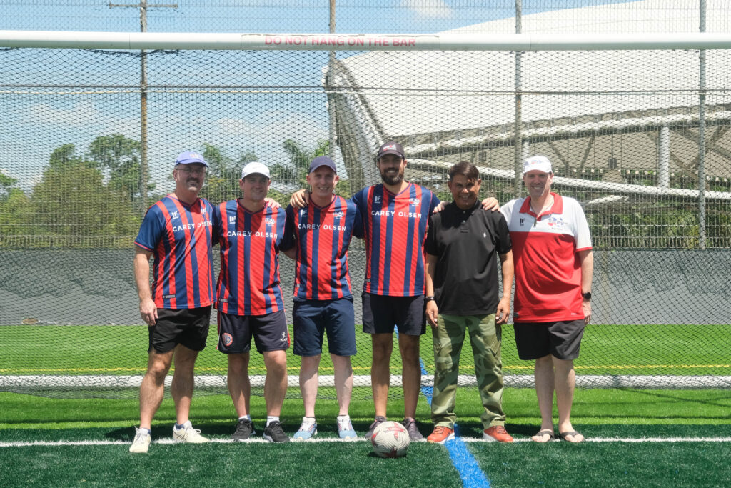 Six ANZA Soccer coaches, some in team jerseys and caps, stand together on an artificial turf soccer field in front of a goal.