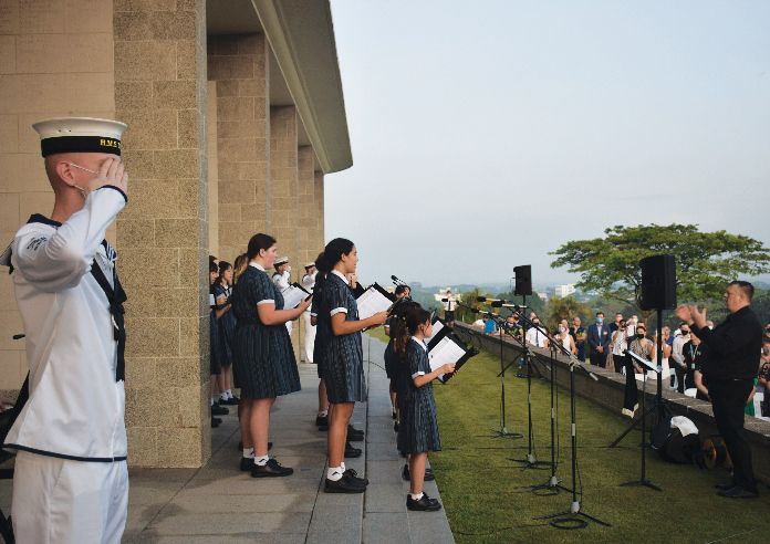 Students singing at ANZAC ceremony, soldiers salute, crowd watches respectfully.