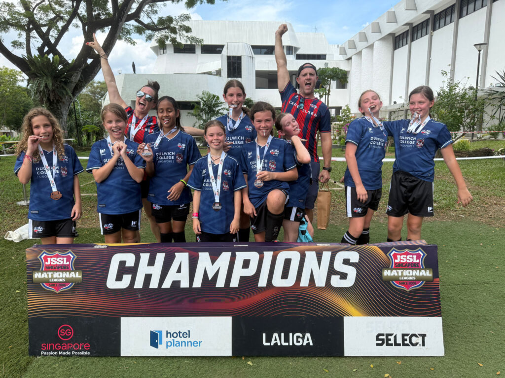 ANZA Soccer girls’ team celebrating with medals after winning the JSSL Singapore National Leagues, posing in front of a ‘Champions’ banner with coaches cheering