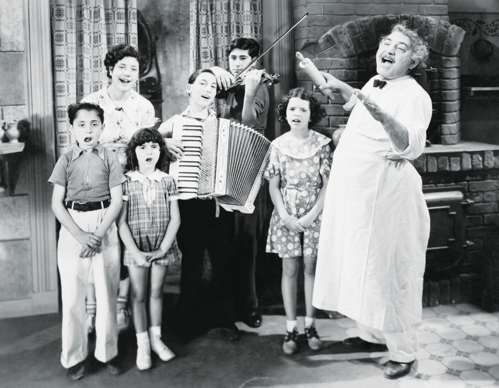 This black-and-white image shows a joyful and nostalgic scene, likely from the mid-20th century. Family joyfully singing and playing music in vintage kitchen setting.